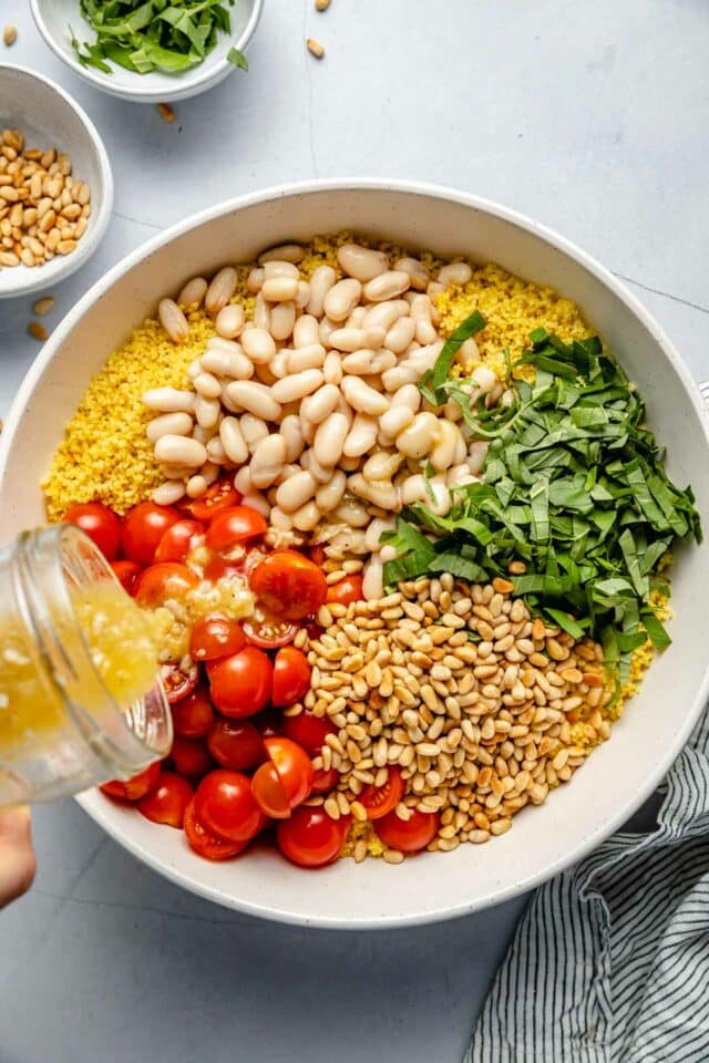 Pouring dressing over couscous, tomatoes, beans, basil and pine nuts. 