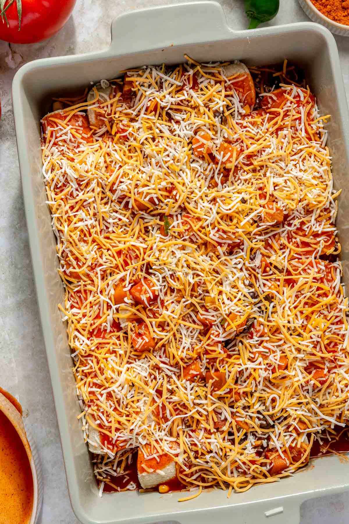 Rolled enchiladas covered with shredded cheese in a pan before baking.