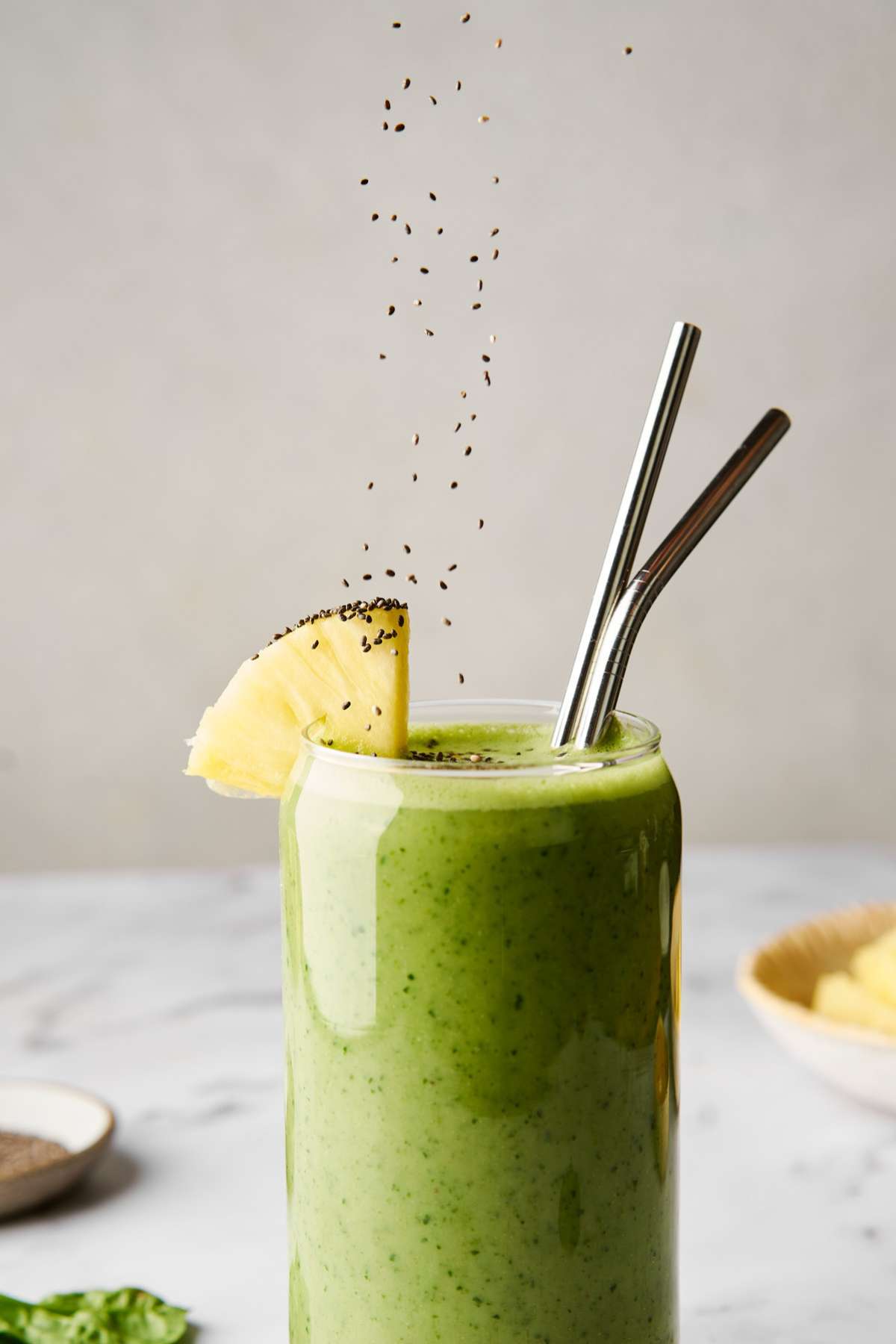 Sprinkling chia seeds over a green smoothie with two metal straws.