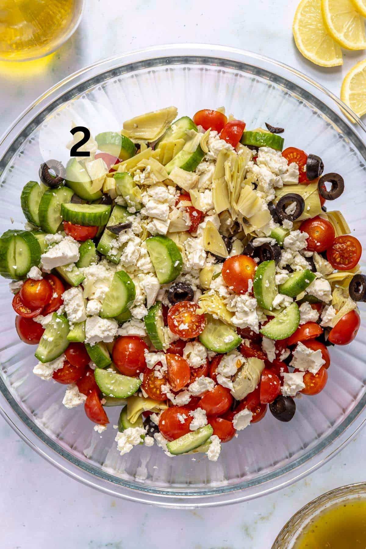 Adding chopped tomatoes, cucumbers, olives and artichoke hearts to a glass bowl.