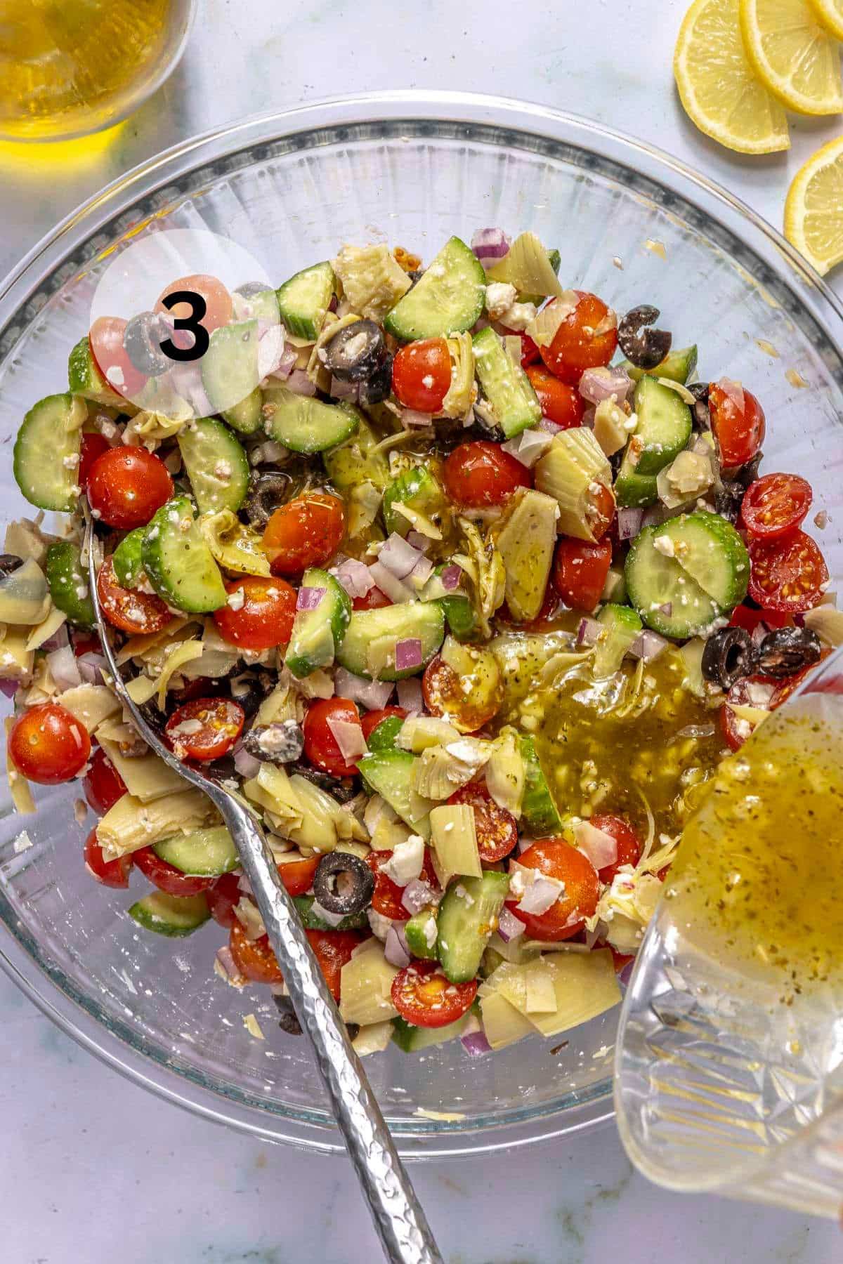 Pouring dressing over a Greek Salad in a glass bowl.