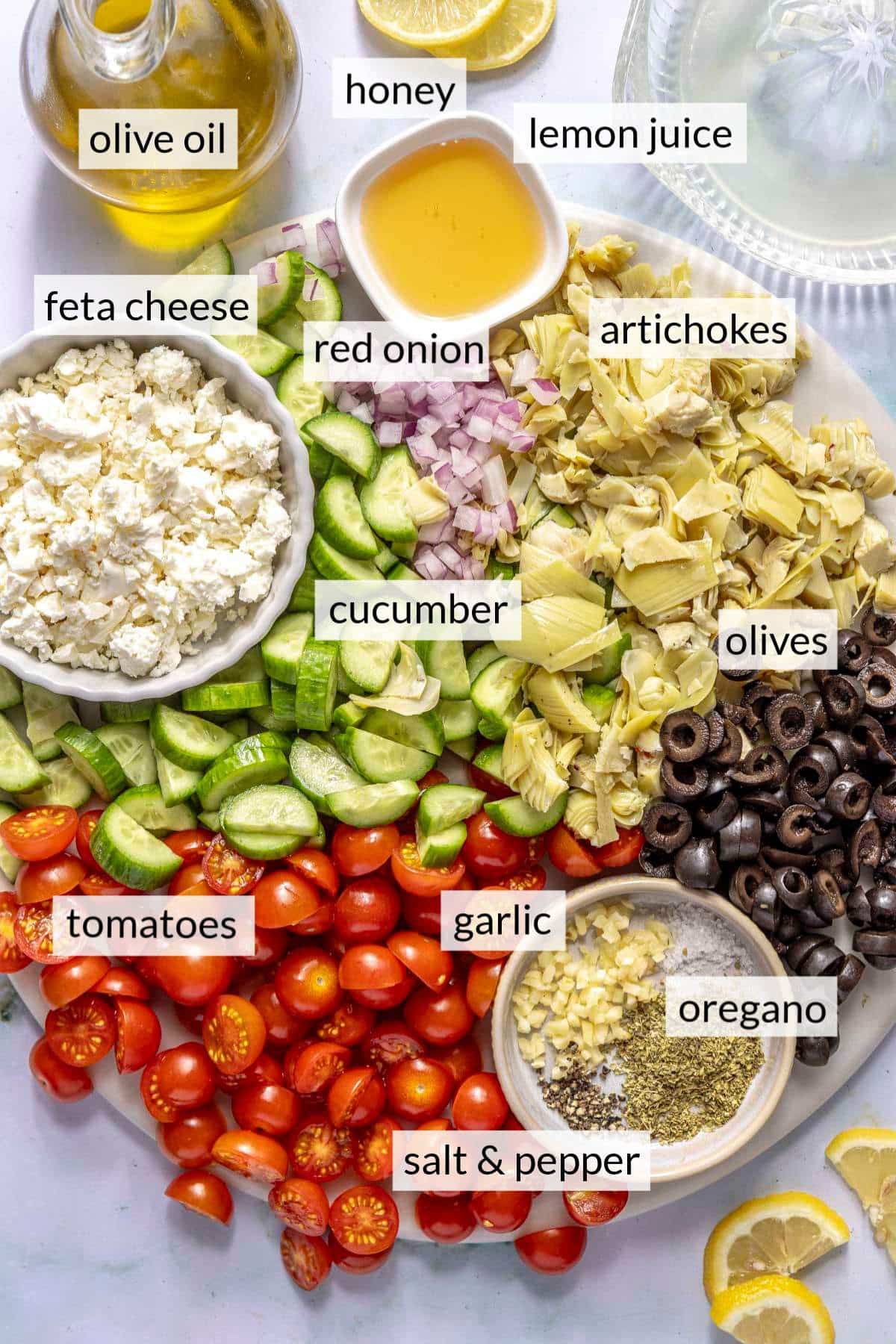 Chopped tomatoes, cucumber, artichokes and black olives on a cutting board near a bowl of seasoning, crumbled feta and honey.