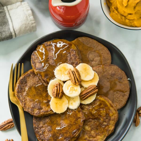 overhead photo of Flourless Pumpkin Pancakes topped with banana slices, pecan halves and maple syrup; served on a black plate with a gold fork and maple syrup on the side.