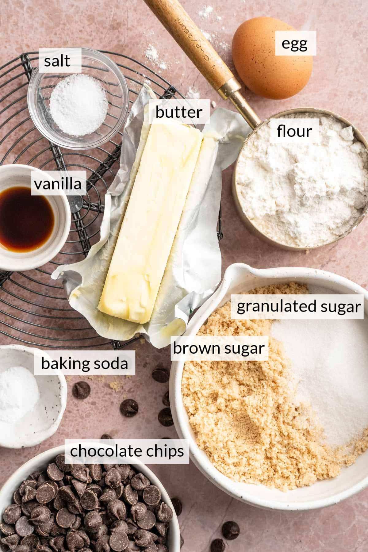 A stick of butter, an egg and bowls of sugar, chocolate chips, flour and vanilla.