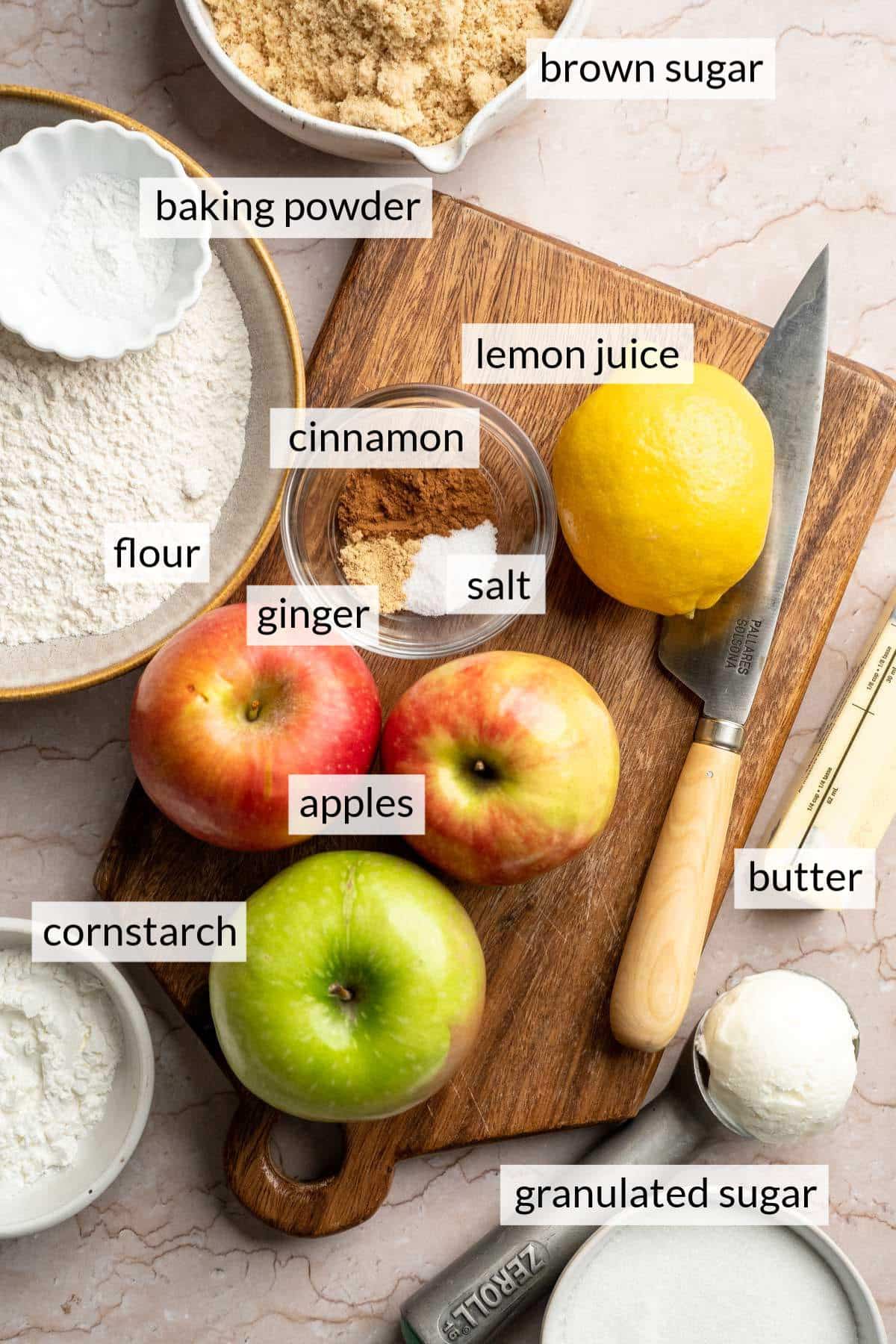 A lemon and three apples on a cutting board near bowls of sugar, flour and spices.