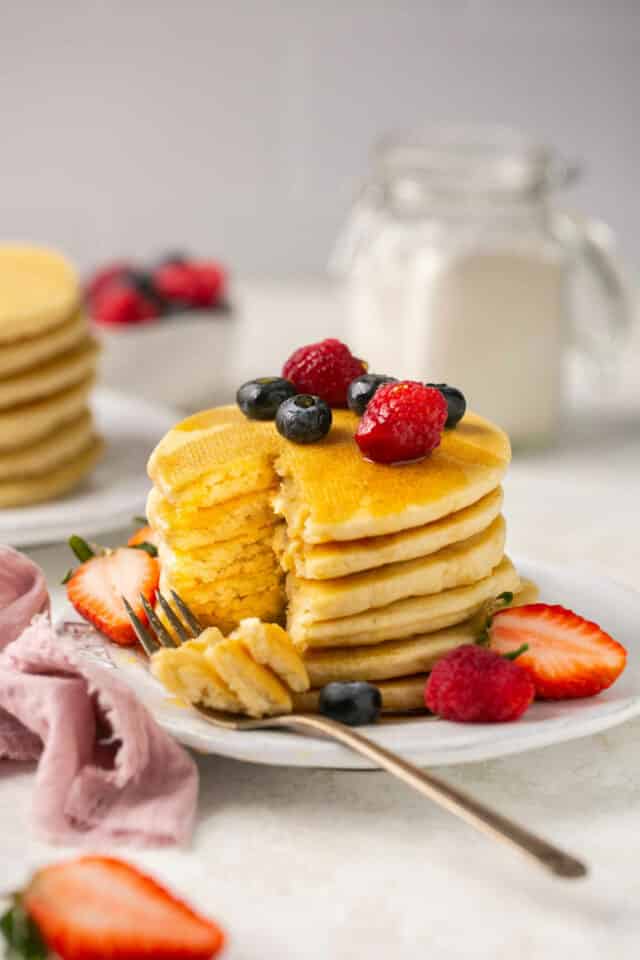 Stack of pancakes on a white plate with berries and maple syrup.