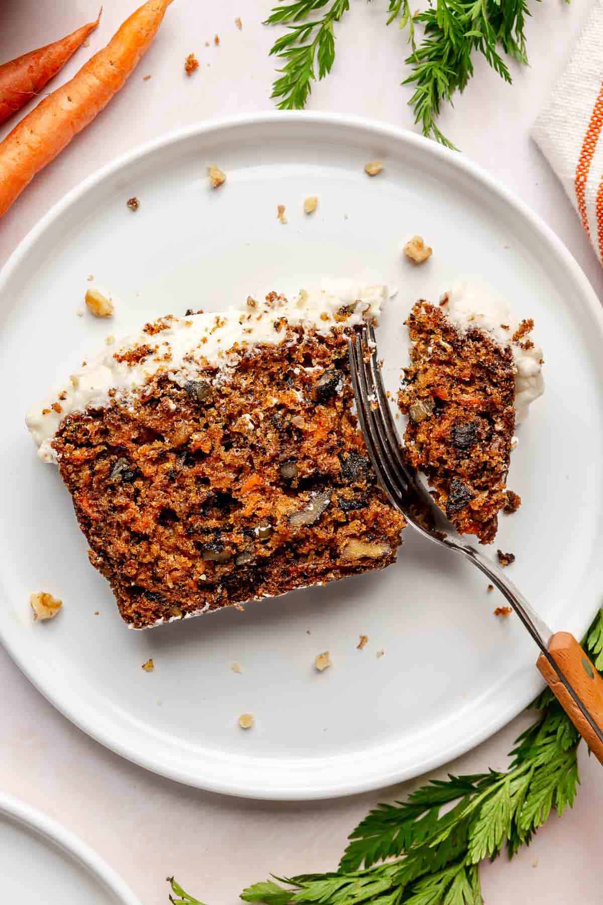 A slice of carrot cake on a plate being cut into with a fork.