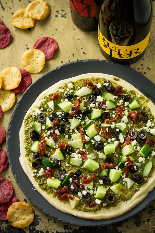 A healthy, tasty, easy Mediterranean layer dip recipe that is a great appetizer or wholesome snack. Loaded with goodies, like hummus, pesto,  feta, olives and sun-dried tomatoes.