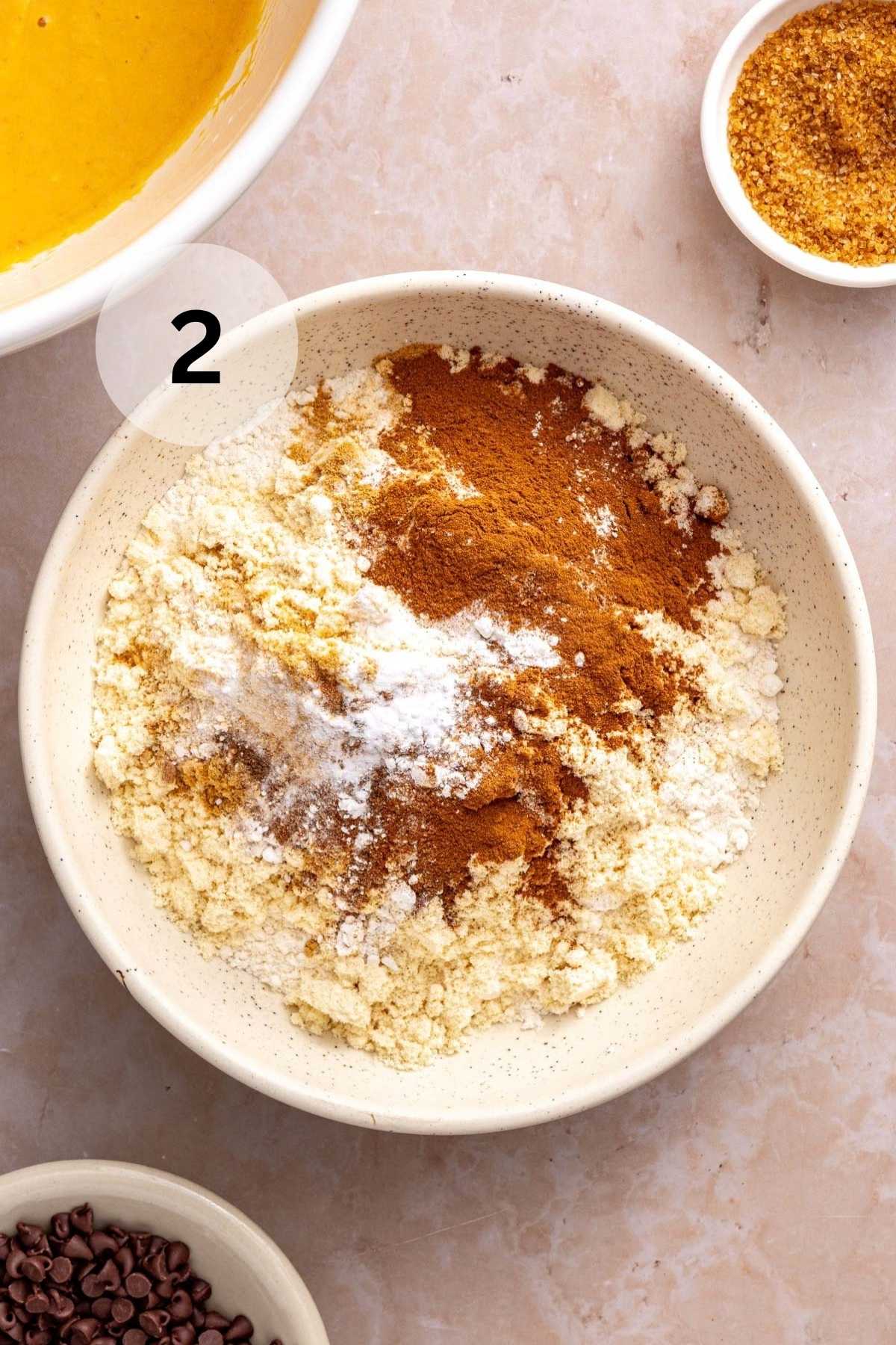 Flour, protein powder, baking powder, baking soda, salt, cinnamon, ginger, nutmeg, and cloves added to a bowl.