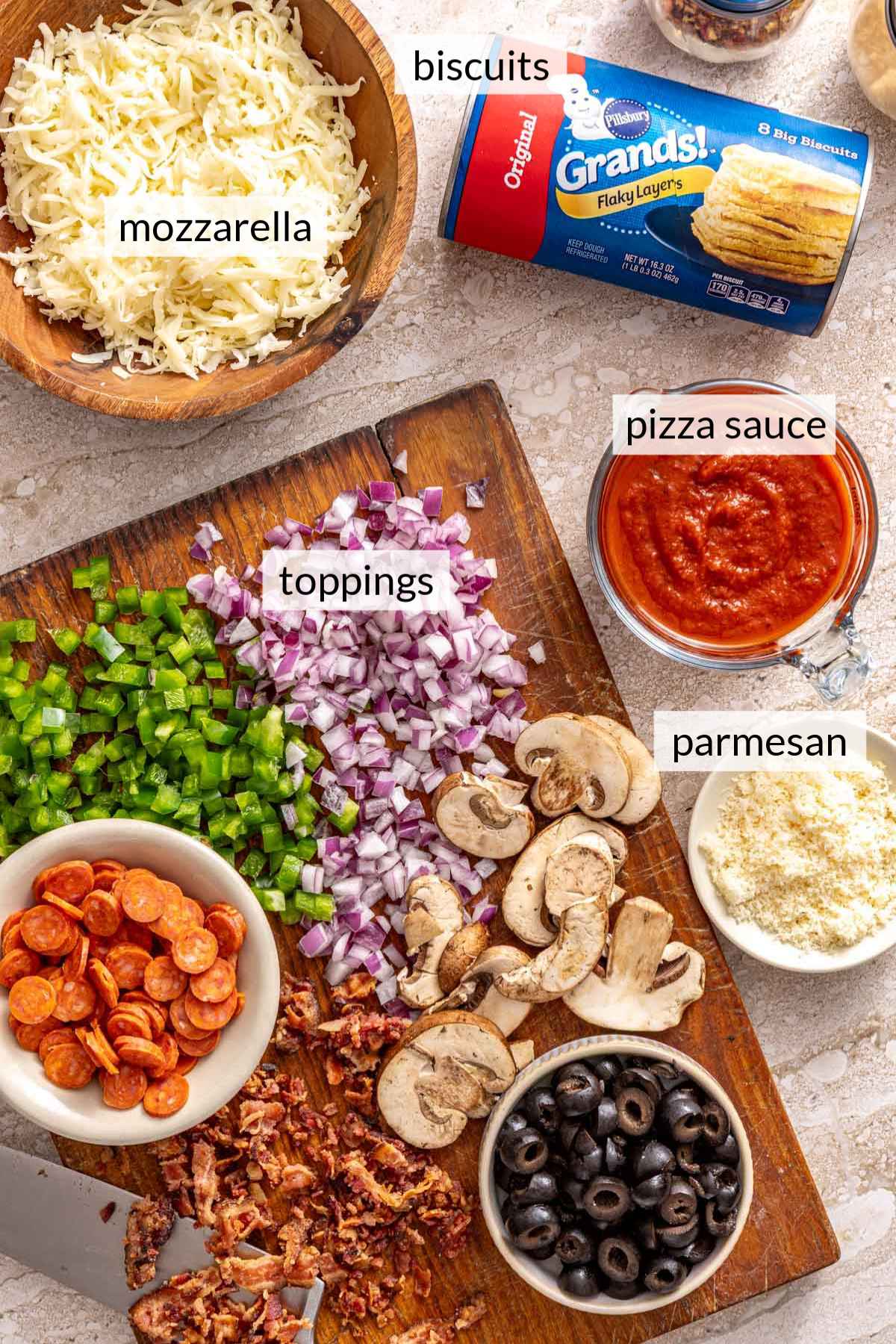 A tube of refrigerated Pillsbury biscuits near shredded cheese, pizza sauce and toppings on a cutting board.