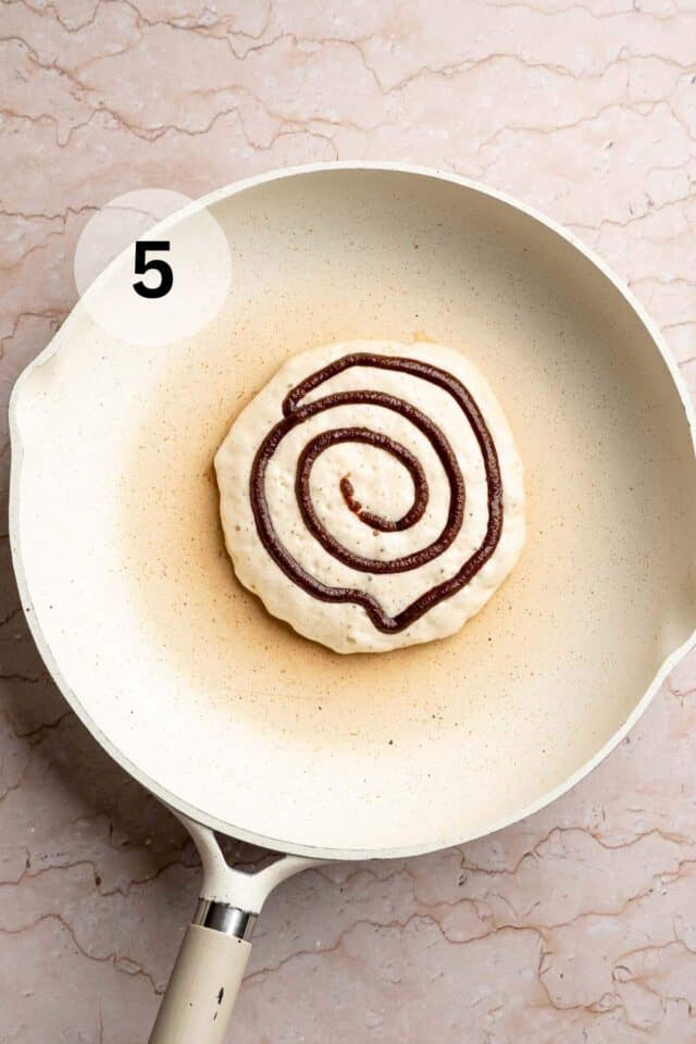 A pancake cooking in a skillet with a cinnamon swirl added to the top.