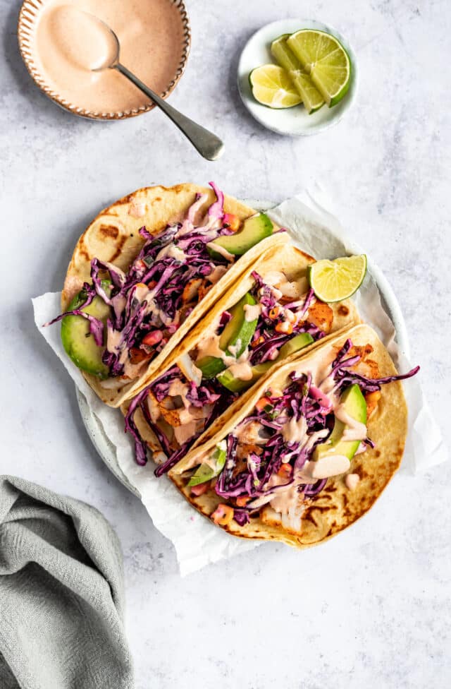 three cod fish tacos topped with slaw, sauce and sliced avocado