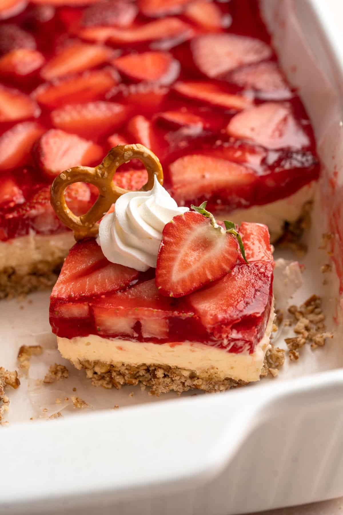 Slice of layered strawberry jello dessert in a pan and topped with whipped cream, a strawberry and pretzel.