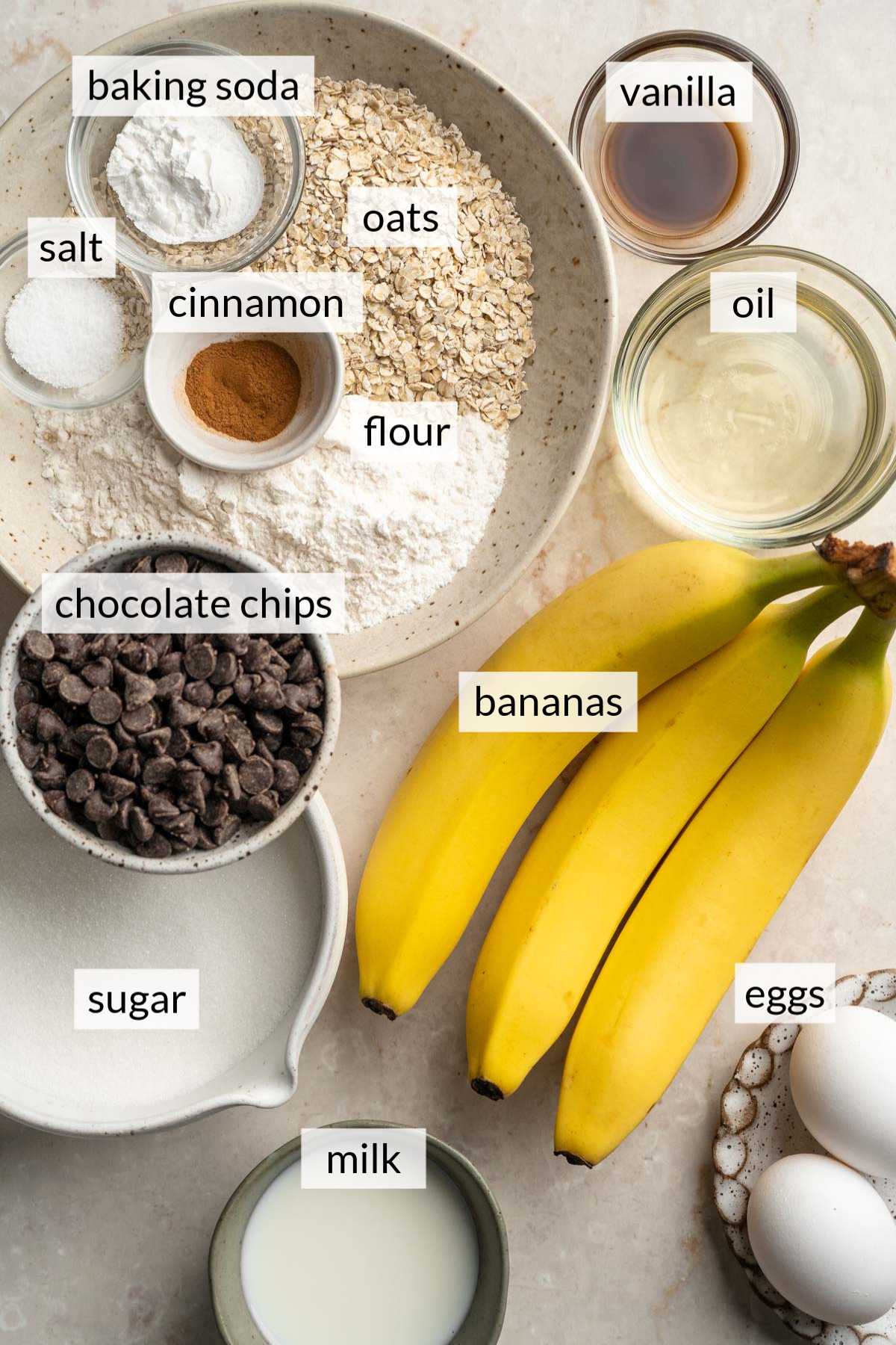 Bananas near eggs and small bowls of oats, vanilla, chocolate chips and sugar.