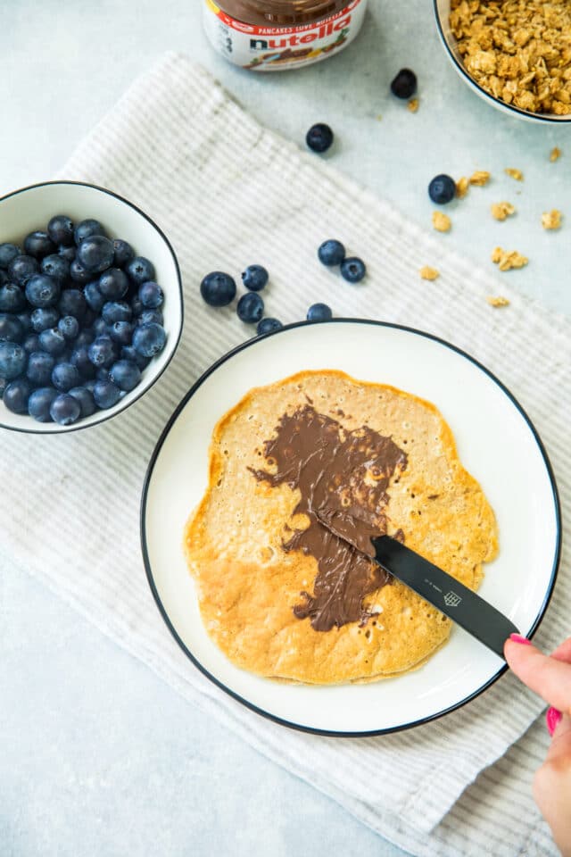 spreading Nutella on a Nutella blueberry protein crepe on a white plate with a black knife. There is a small white bowl of blueberries on the side.