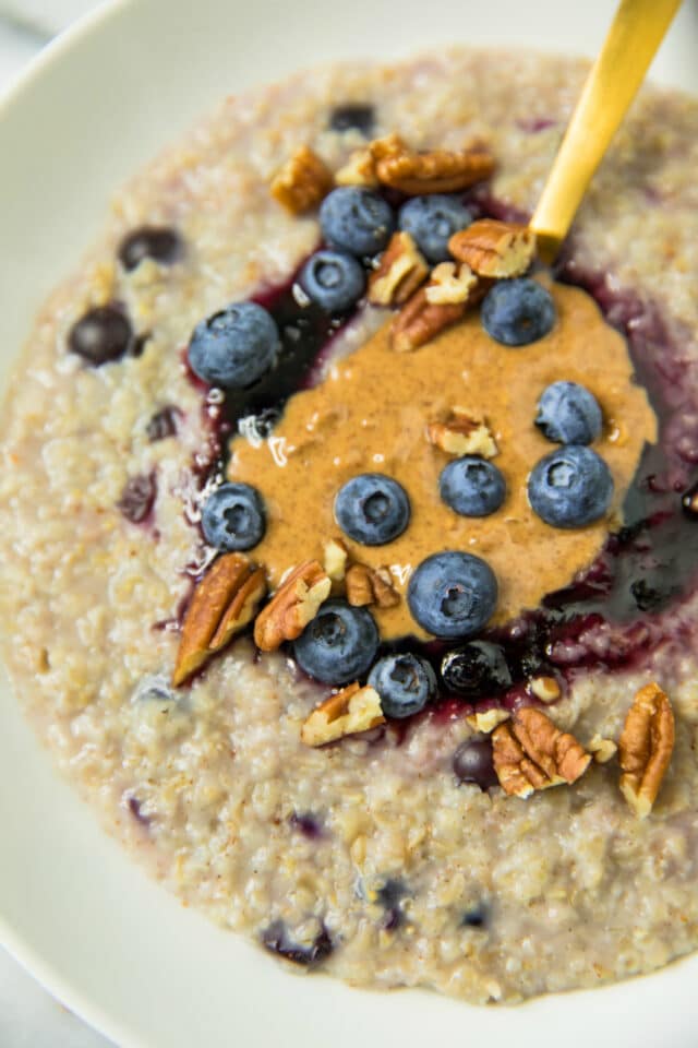 closeup view of oatmeal in a white bowl topped with blueberries, almond butter, pecans and blueberry jam