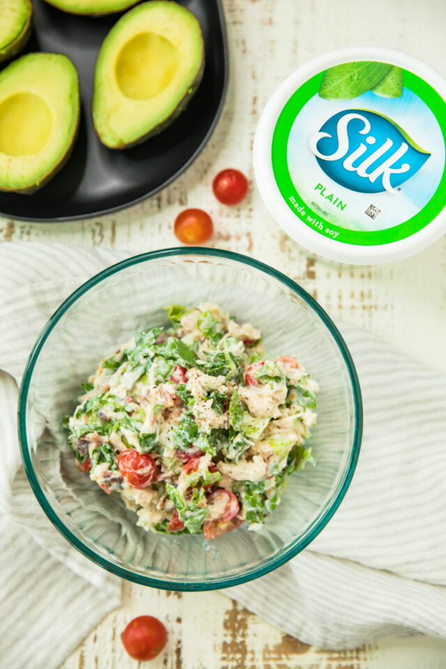 The chicken salad for the BLT Chicken Salad Stuffed Avocados in a glass bowl, avocado halves on a black plate and Silk Plain Yogurt container
