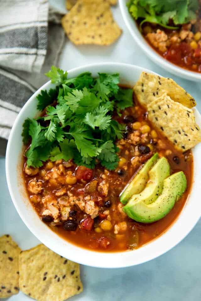 closeup overhead of healthy taco soup served in a white bowl topped with avocado slices, chips and cilantro