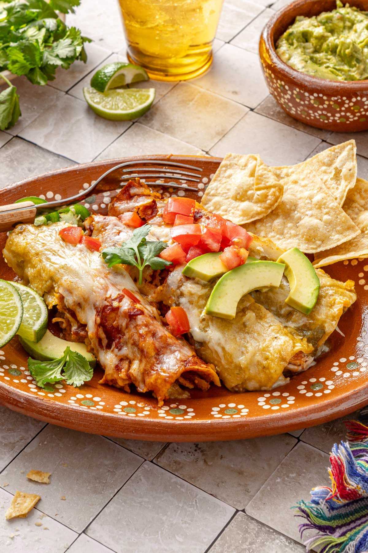 Two chicken enchiladas topped with diced tomatoes and avocado slices on a plate with chips.