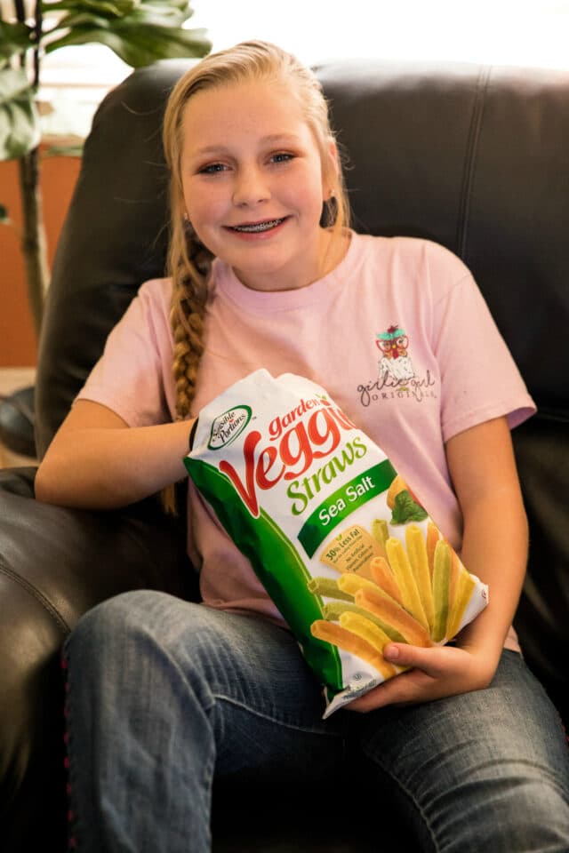 young girl, smiling, with hand in bag of garden veggie straws