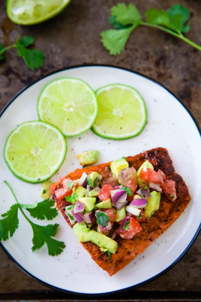 Salmon with avocado topping on a white plate with lime slices and fresh cilantro on the side