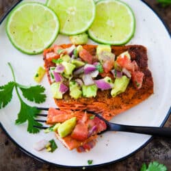 Pan Seared Salmon with Avocado Salsa on a white plate with a black fork taking a bite
