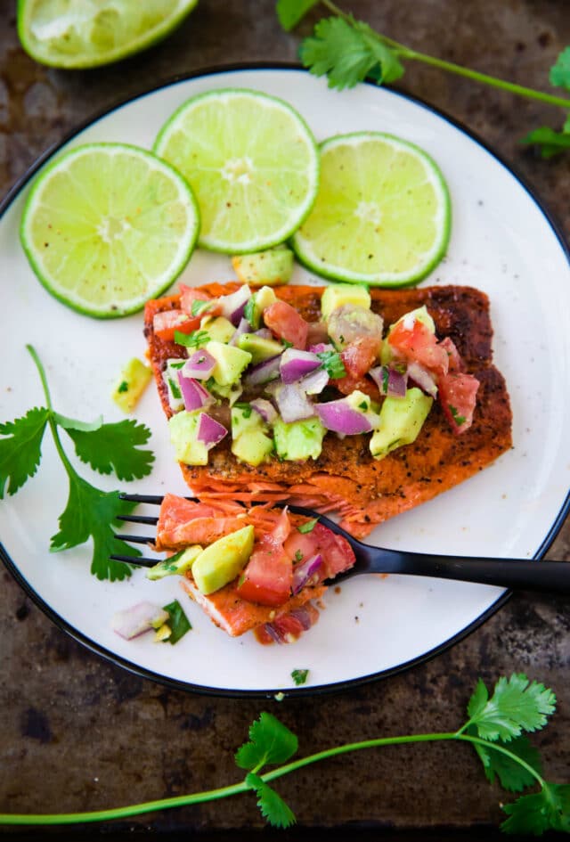 Pan Seared Salmon with Avocado Salsa on a white plate with a black fork taking a bite 