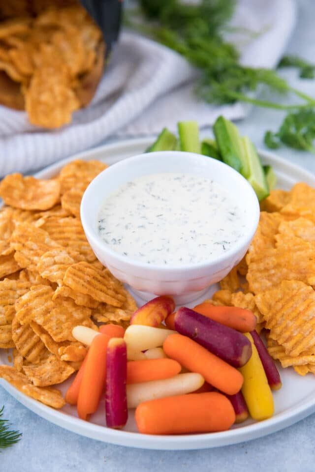 Homemade Ranch Dip in a white bowl on a platter with chips, baby carrots and cucumber sticks
