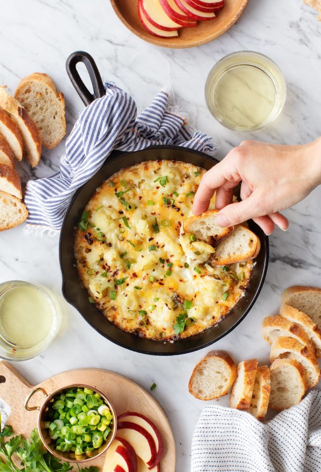 cheesy dip in a cast iron skillet with a woman's hand dipping bread into it
