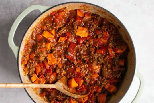 Using a wooden spoon to stir tomatoes with ground beef in a large pot.