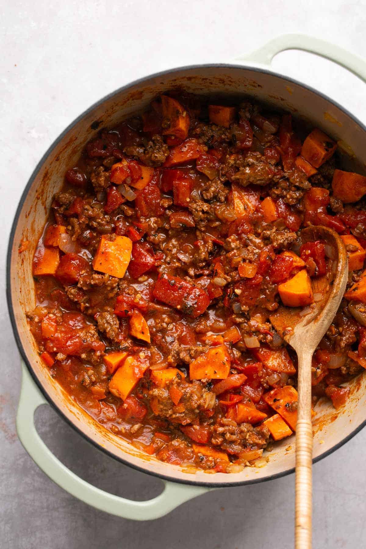 Using a wooden spoon to stir tomatoes with ground beef in a large pot.