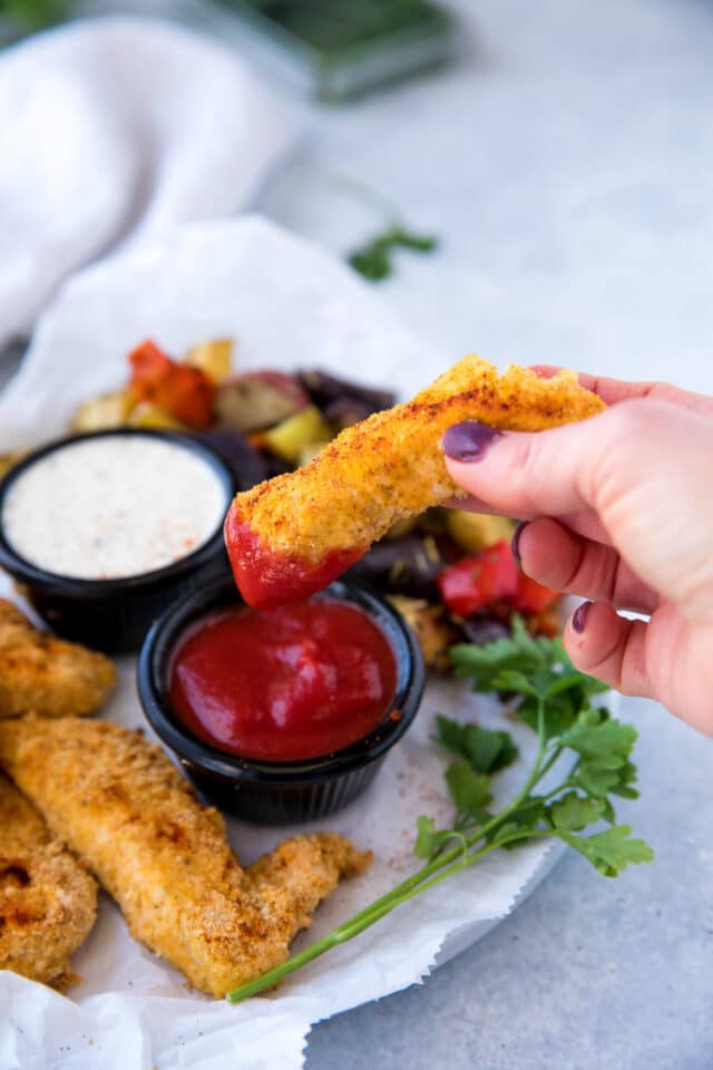 woman's hand holding a chicken tender and dipping it into ketchup 