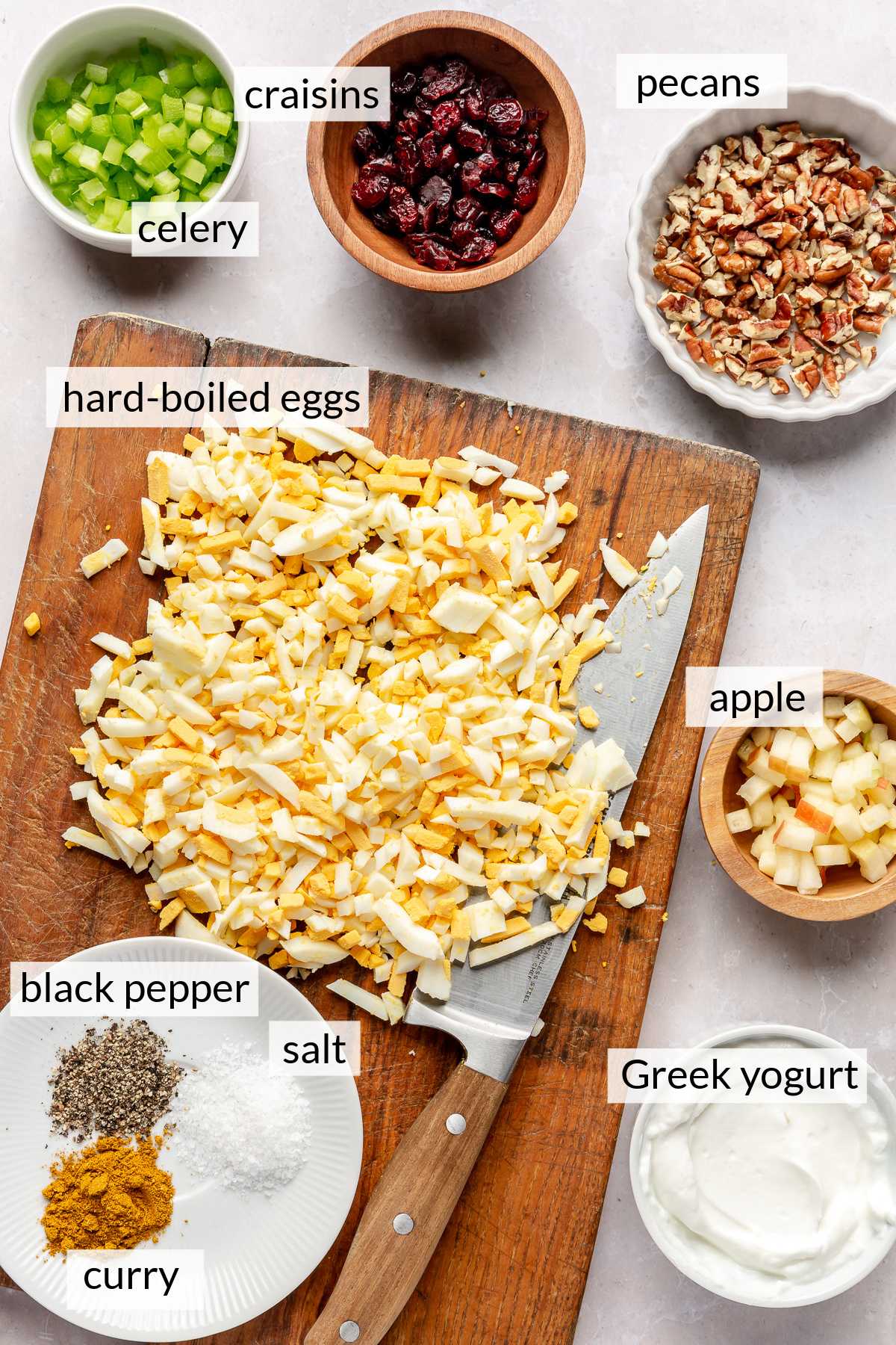 Chopped hard-boiled eggs on a cutting board near bowls of chopped pecans, craisins, celery and greek yogurt.
