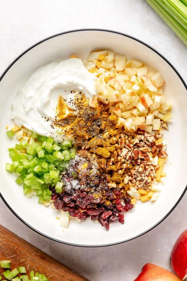 Greek yogurt added to a bowl with diced apple, celery, boiled eggs, and raisins.