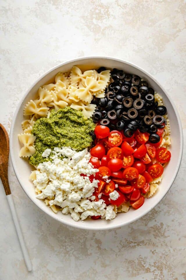 Combining short pasta with black olives, tomatoes, feta cheese and pesto in a large bowl.