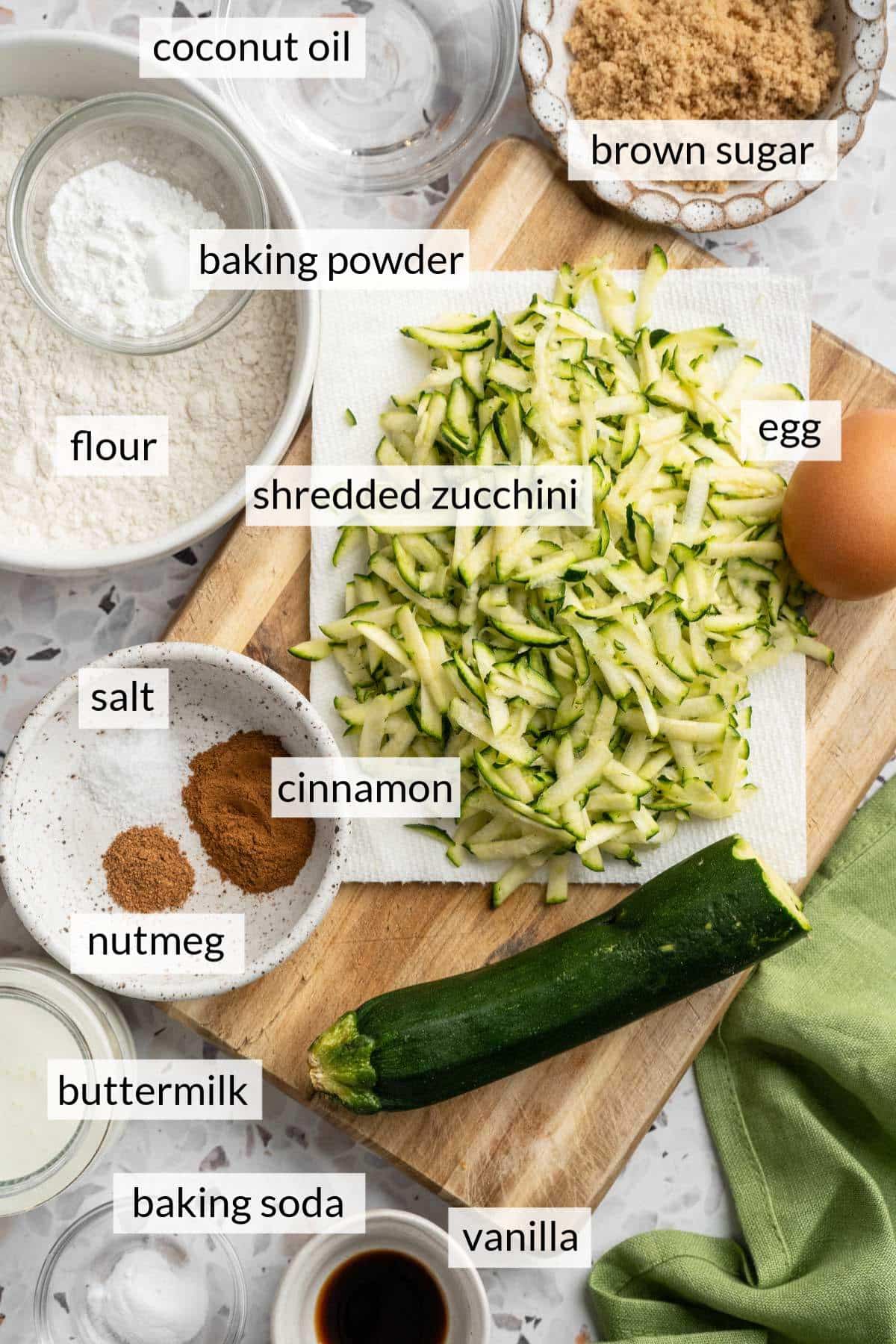 Shredded zucchini near small bowls of sugar, flour, spices, vanilla and buttermilk.