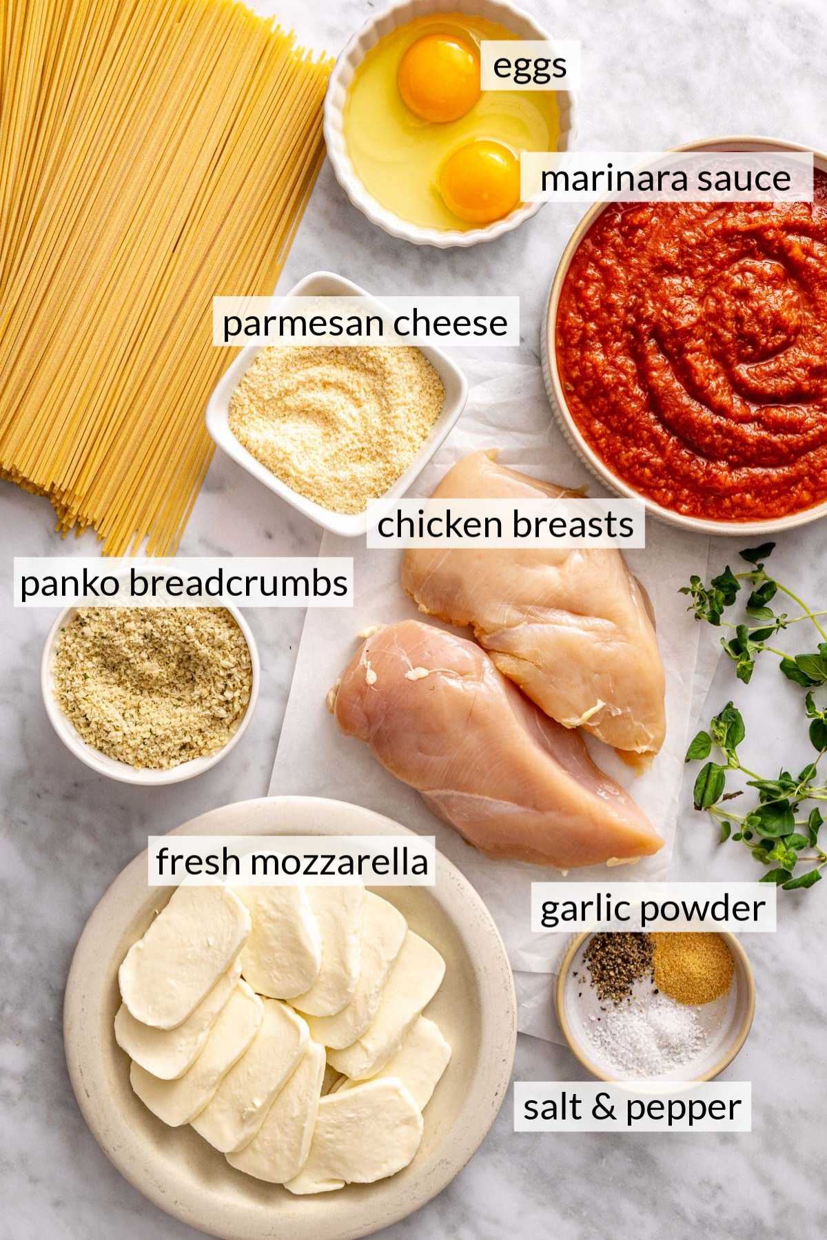 Raw chicken breasts near small bowls with marinara sauce, breadcrumbs, parmesan cheese and mozzarella.