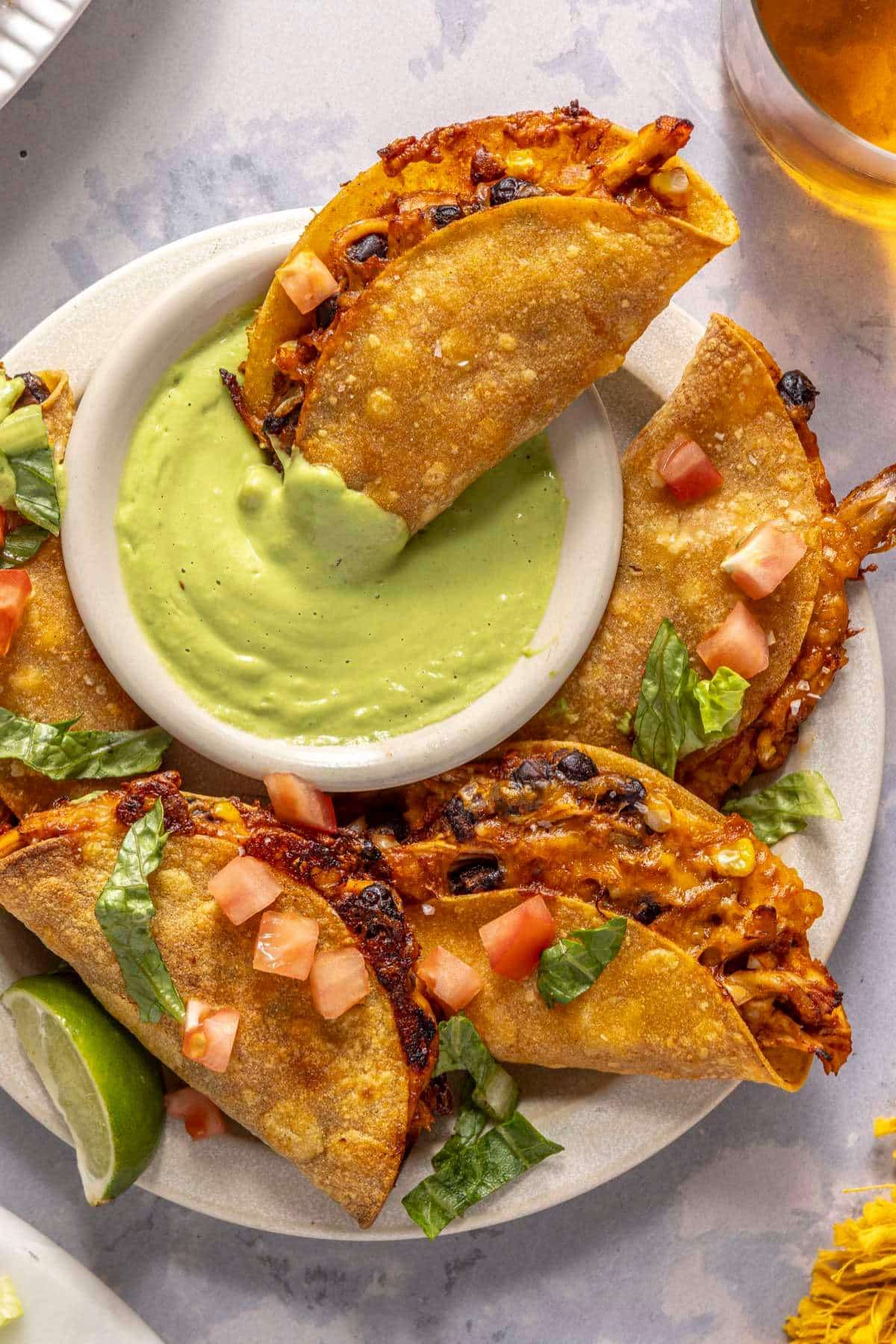 Crispy chicken tacos served with a green sauce and one dipped in.