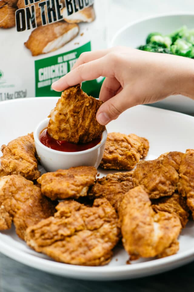 child's hand dipping a Healthy Baked Chicken Tenders into ketchup