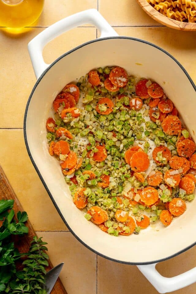 Carrots, onions and celery cooking with seasonings in a large white pot.