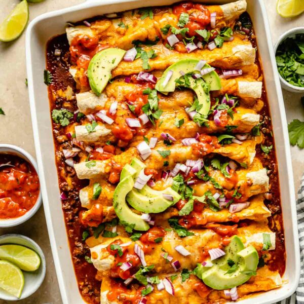 Beef enchiladas in a white casserole dish topped with avocado slices and fresh cilantro.