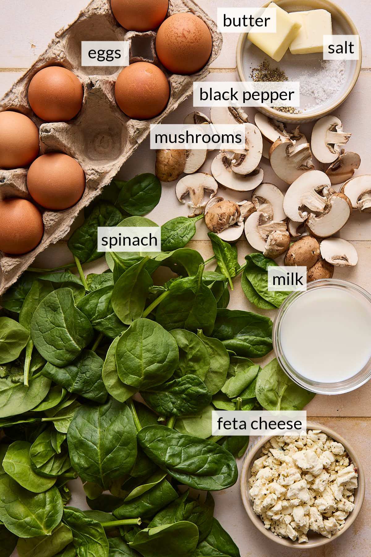 Fresh baby spinach near a carton of eggs, sliced mushrooms and small cups of milk and feta cheese crumbles.