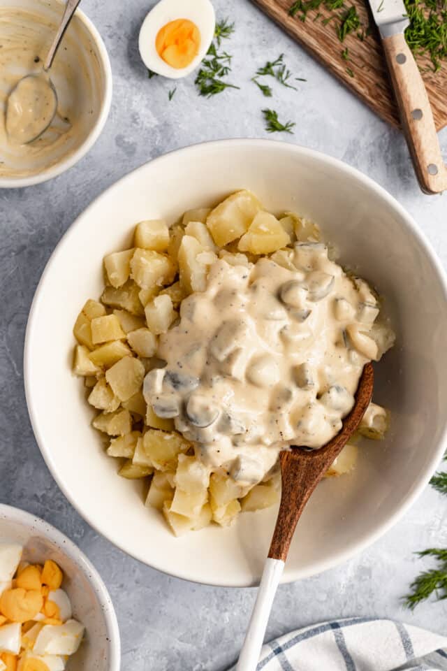 adding potato salad sauce to cooked potatoes in a white bowl 
