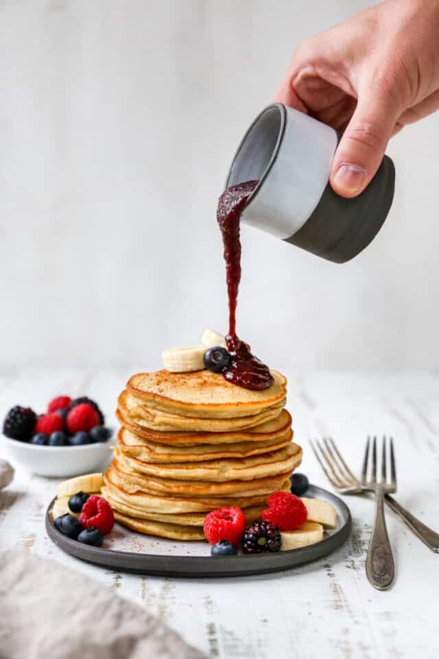 pouring raspberry syrup over the top of a stack of pancakes