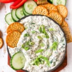 spinach dip in a bowl with crackers and cucumbers dipped in
