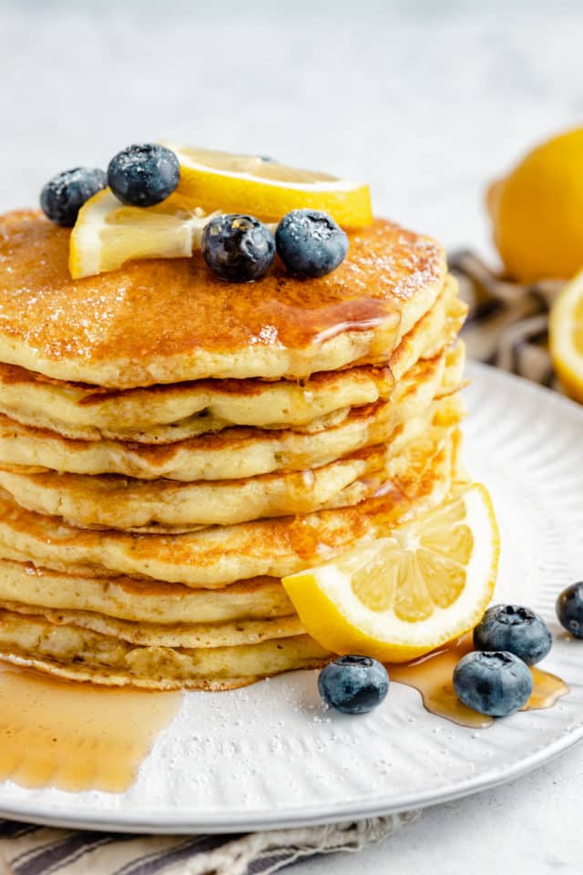 stack of lemon ricotta pancakes topped with blueberries, syrup and sliced lemon