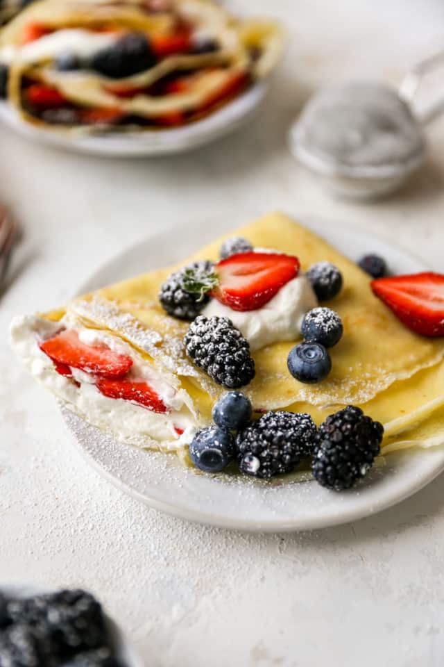homemade crepe served on a small white plate with berries and a dusting of powdered sugar