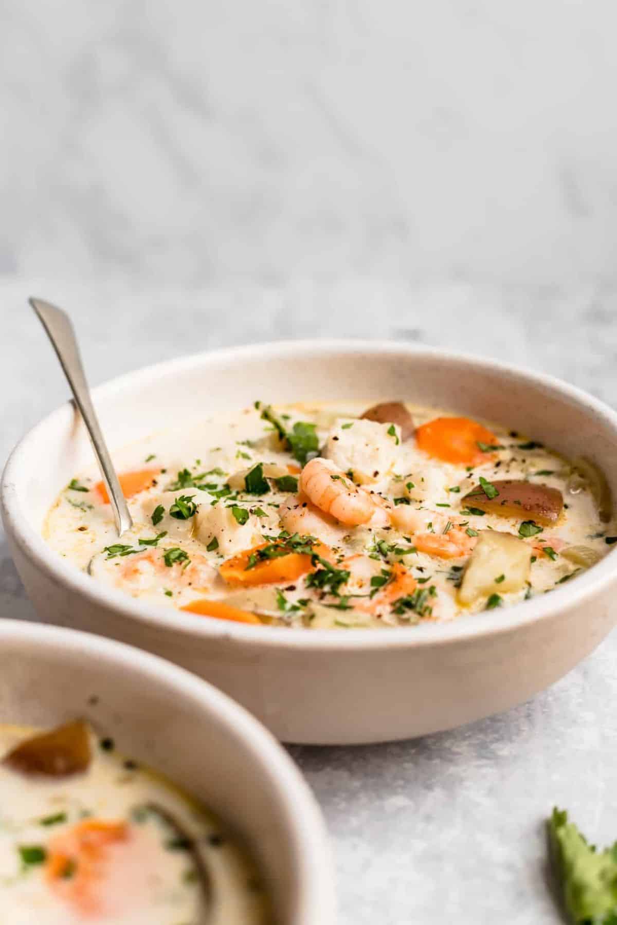 Seafood chowder with shrimp, fish, potatoes and carrots in a white bowl with a spoon.