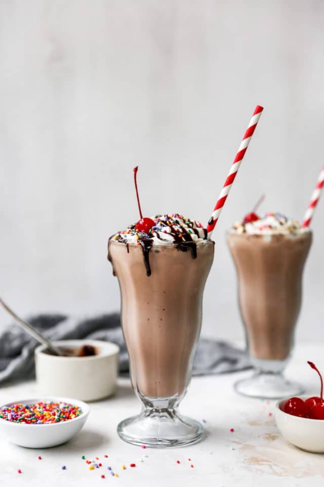 a large glass with a chocolate milkshake topped with whipped cream, sprinkles and a cherry 