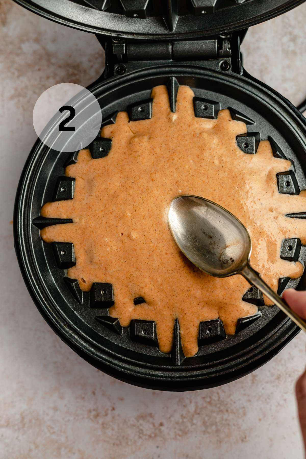 Spreading waffle batter over waffle iron with the back of a spoon.